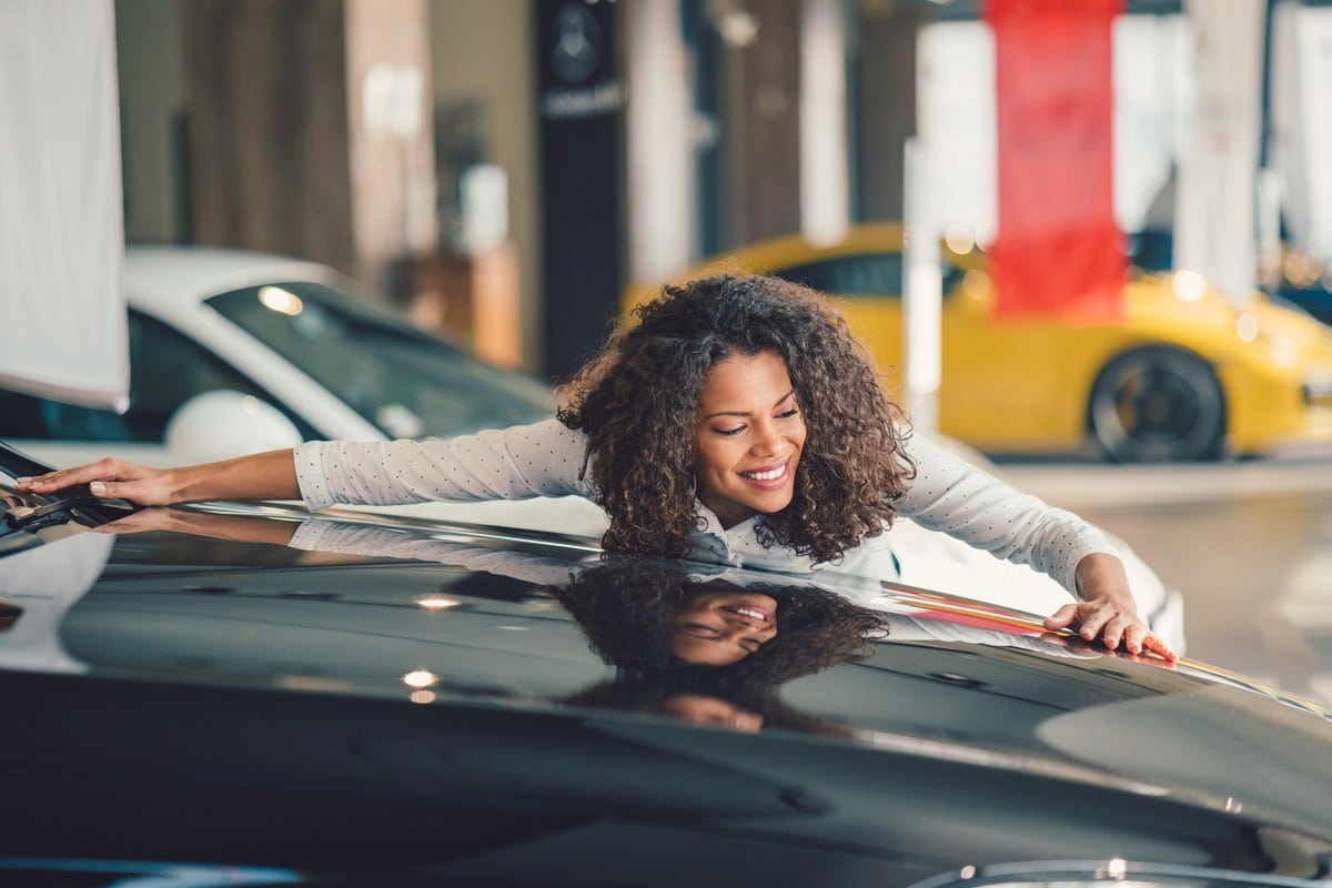 Uma mulher negra, que acabou de financiar o primeiro carro. Ela está abraçada a um carro preto, dentro da concessionária.